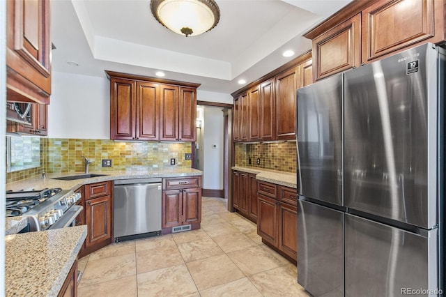 kitchen with a tray ceiling, backsplash, appliances with stainless steel finishes, a sink, and light stone countertops