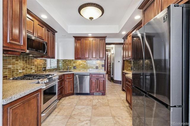 kitchen with a raised ceiling, light stone counters, a sink, stainless steel appliances, and backsplash