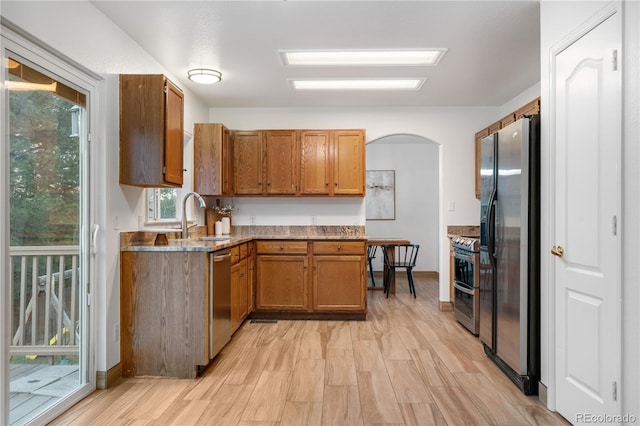 kitchen featuring stainless steel appliances, arched walkways, a sink, brown cabinets, and light wood finished floors