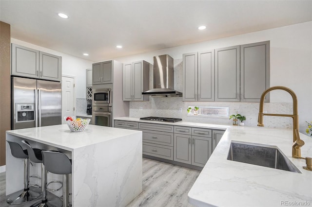 kitchen with light stone counters, gray cabinets, appliances with stainless steel finishes, wall chimney exhaust hood, and a sink