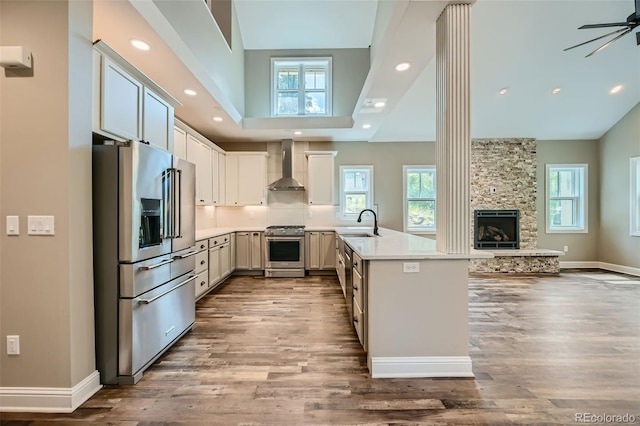 kitchen with stainless steel appliances, kitchen peninsula, ceiling fan, a stone fireplace, and wall chimney range hood