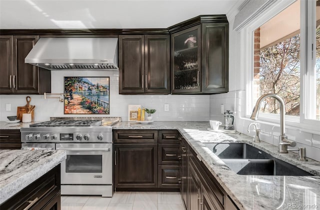 kitchen featuring stainless steel stove, a sink, wall chimney range hood, tasteful backsplash, and glass insert cabinets