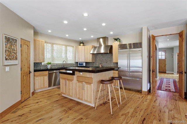 kitchen featuring light brown cabinetry, a kitchen island, dark countertops, stainless steel appliances, and wall chimney range hood