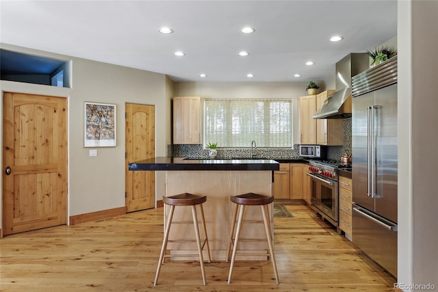 kitchen with a breakfast bar, light brown cabinetry, dark countertops, wall chimney range hood, and high quality appliances