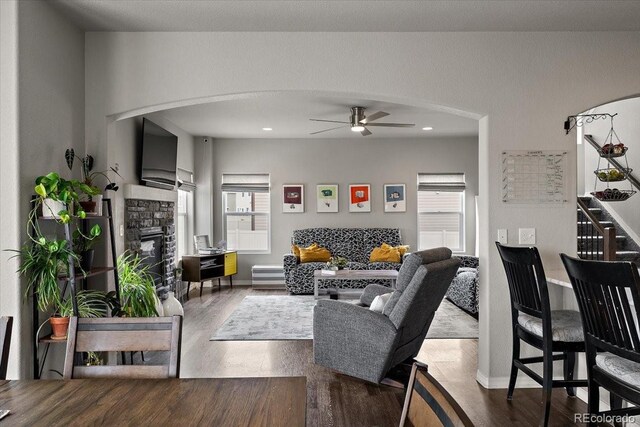 living room with wood finished floors, a ceiling fan, a stone fireplace, and arched walkways