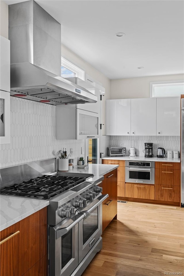 kitchen with tasteful backsplash, appliances with stainless steel finishes, light wood-style floors, modern cabinets, and wall chimney exhaust hood