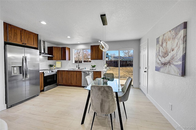 kitchen featuring wall chimney range hood, appliances with stainless steel finishes, pendant lighting, and light hardwood / wood-style floors