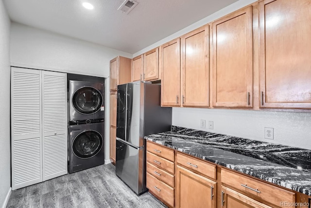 kitchen with light brown cabinets, dark stone countertops, stainless steel fridge, hardwood / wood-style flooring, and stacked washer / dryer