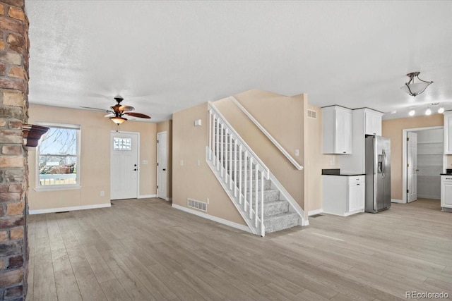 unfurnished living room with light wood-style flooring, a ceiling fan, visible vents, stairs, and baseboards