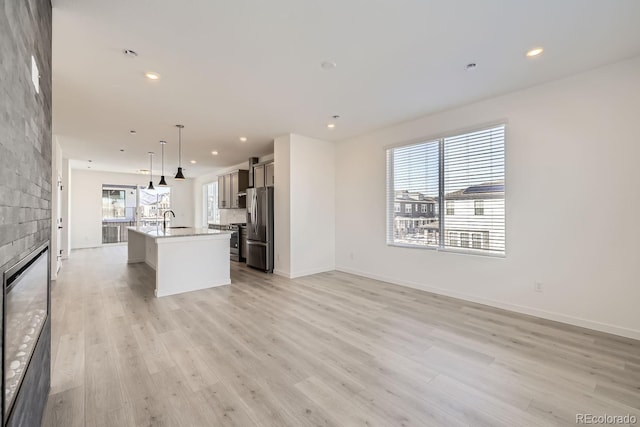 kitchen with light hardwood / wood-style flooring, appliances with stainless steel finishes, a large fireplace, an island with sink, and decorative light fixtures