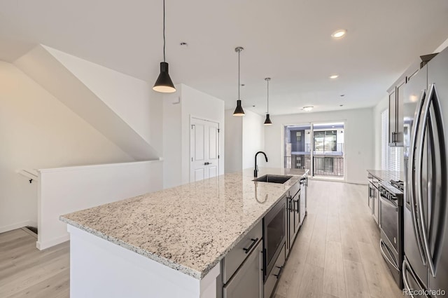 kitchen with sink, light stone counters, appliances with stainless steel finishes, an island with sink, and pendant lighting