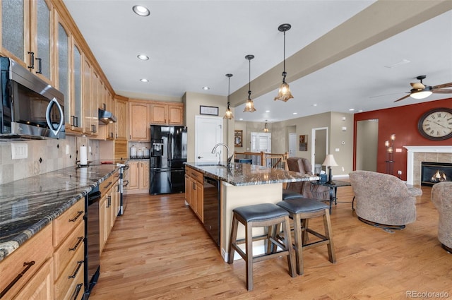 kitchen with black appliances, a fireplace, dark stone countertops, light hardwood / wood-style flooring, and a center island with sink