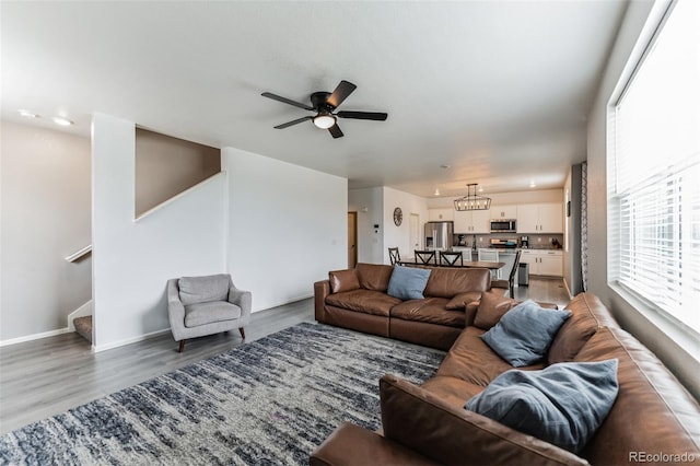 living room with stairs, ceiling fan with notable chandelier, light wood-style flooring, and baseboards