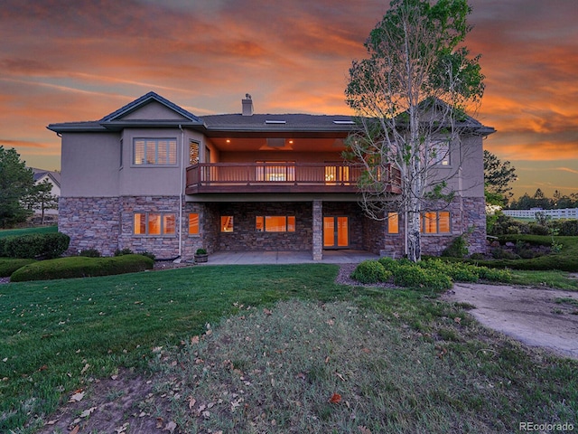 back of house at dusk with stone siding, a patio area, a lawn, a chimney, and a deck