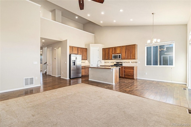 kitchen with ceiling fan with notable chandelier, appliances with stainless steel finishes, dark colored carpet, an island with sink, and decorative light fixtures