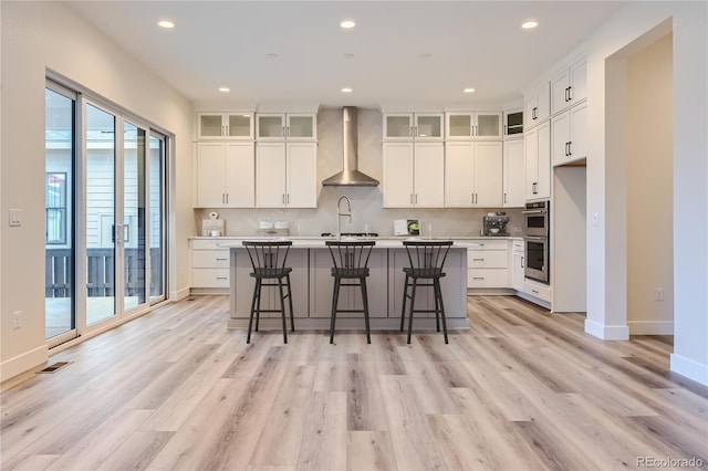 kitchen with a breakfast bar, white cabinets, light hardwood / wood-style flooring, wall chimney exhaust hood, and an island with sink