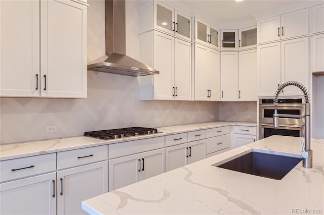 kitchen featuring light stone countertops, wall chimney exhaust hood, multiple ovens, white cabinets, and stainless steel gas stovetop