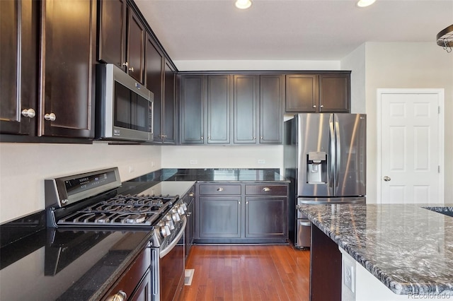 kitchen with appliances with stainless steel finishes, dark hardwood / wood-style flooring, dark brown cabinets, and dark stone counters