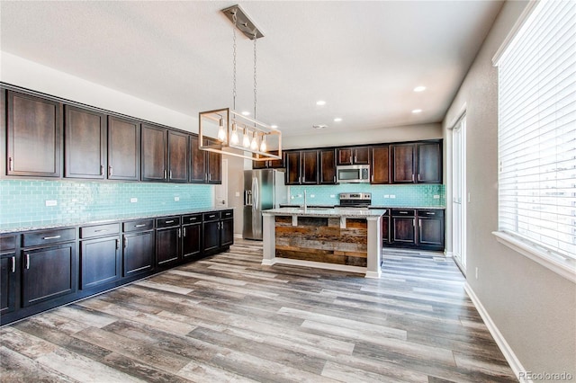 kitchen with appliances with stainless steel finishes, pendant lighting, an island with sink, light stone counters, and dark brown cabinetry