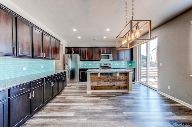 kitchen featuring pendant lighting, appliances with stainless steel finishes, dark brown cabinets, light stone counters, and an island with sink