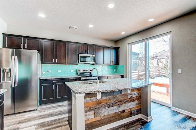 kitchen with appliances with stainless steel finishes, light stone countertops, an island with sink, and backsplash