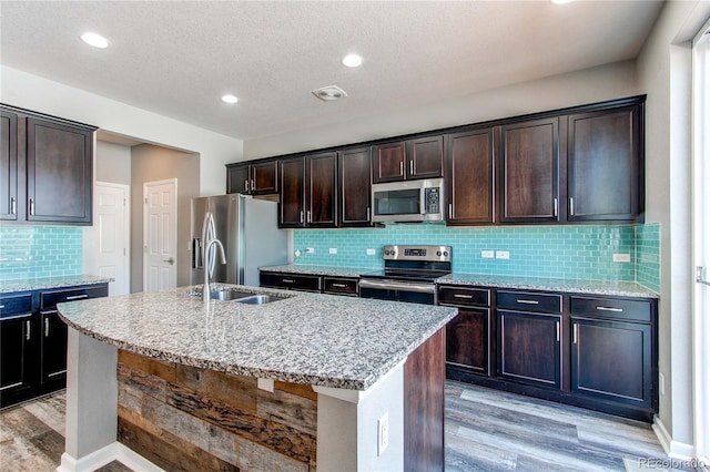 kitchen featuring sink, dark brown cabinets, stainless steel appliances, light stone counters, and an island with sink