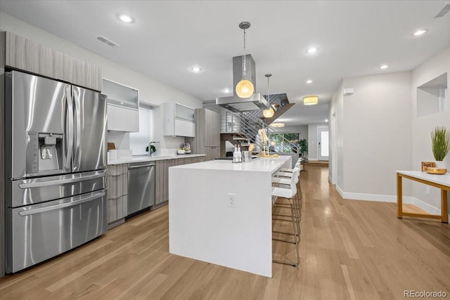 kitchen featuring appliances with stainless steel finishes, island range hood, decorative light fixtures, a center island with sink, and light hardwood / wood-style flooring