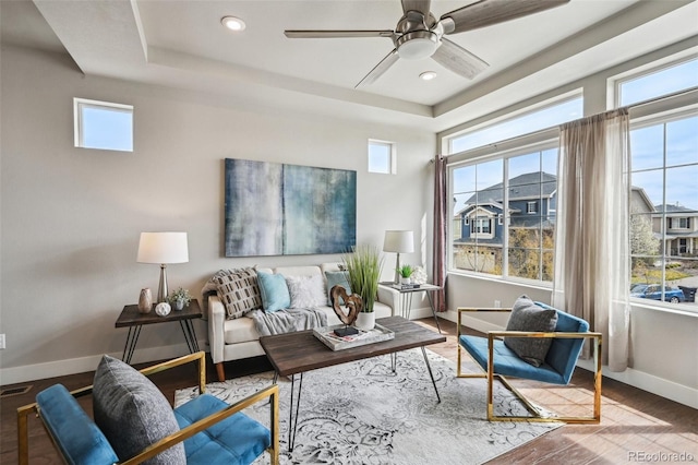 living area with wood finished floors, recessed lighting, a ceiling fan, a tray ceiling, and baseboards