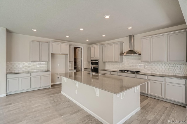 kitchen with wall chimney range hood, an island with sink, light wood-style flooring, stainless steel appliances, and a sink