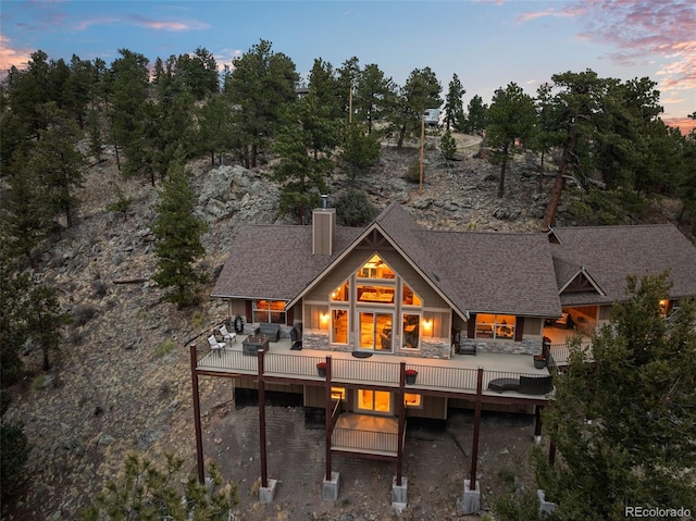 back of property at dusk with a chimney, a shingled roof, stone siding, and a deck
