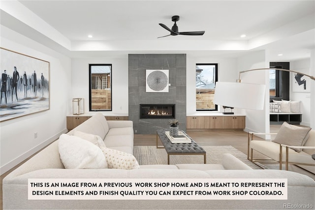 living room featuring a raised ceiling, a tiled fireplace, ceiling fan, and light hardwood / wood-style flooring