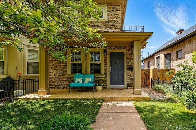 doorway to property featuring stone siding, fence, a balcony, and a porch