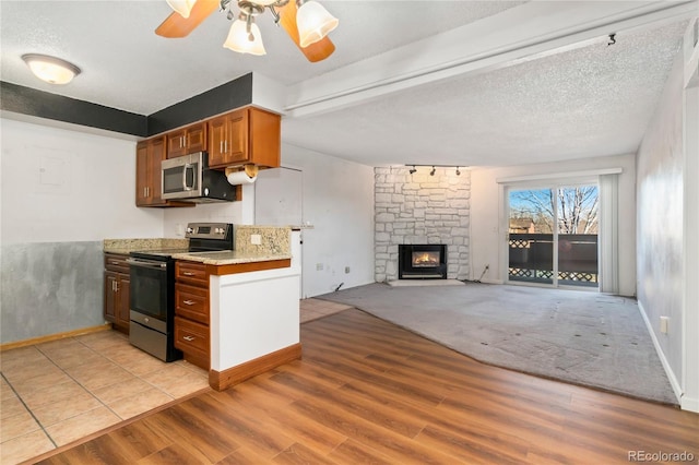 kitchen featuring brown cabinets, stainless steel appliances, light wood-style floors, a stone fireplace, and ceiling fan