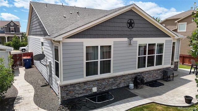 back of house with stone siding, roof with shingles, a patio area, and fence