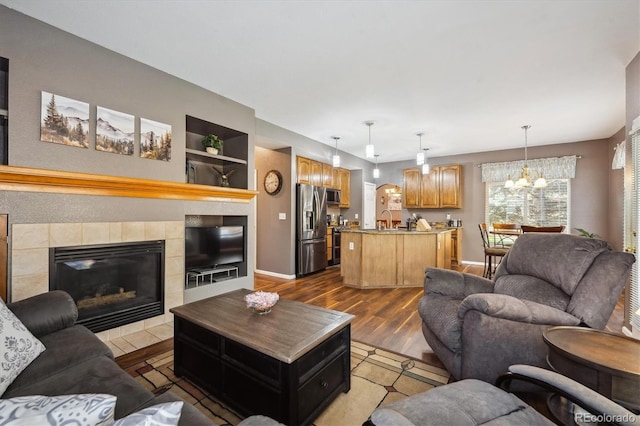 living room featuring an inviting chandelier, built in features, baseboards, dark wood-style floors, and a tile fireplace
