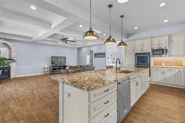 kitchen with sink, hanging light fixtures, appliances with stainless steel finishes, light stone countertops, and a kitchen island with sink