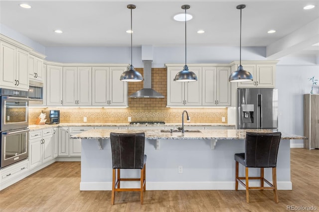 kitchen featuring pendant lighting, appliances with stainless steel finishes, light stone counters, a center island with sink, and wall chimney exhaust hood