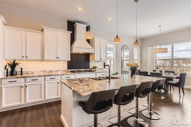 kitchen with custom exhaust hood, white cabinetry, a kitchen island with sink, decorative backsplash, and sink