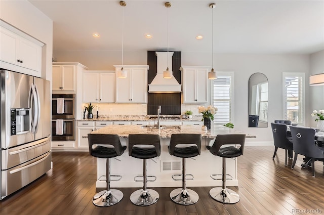 kitchen with stainless steel appliances, custom exhaust hood, decorative backsplash, an island with sink, and dark wood finished floors