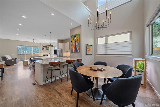 dining room with light wood-type flooring, ceiling fan with notable chandelier, a towering ceiling, and sink