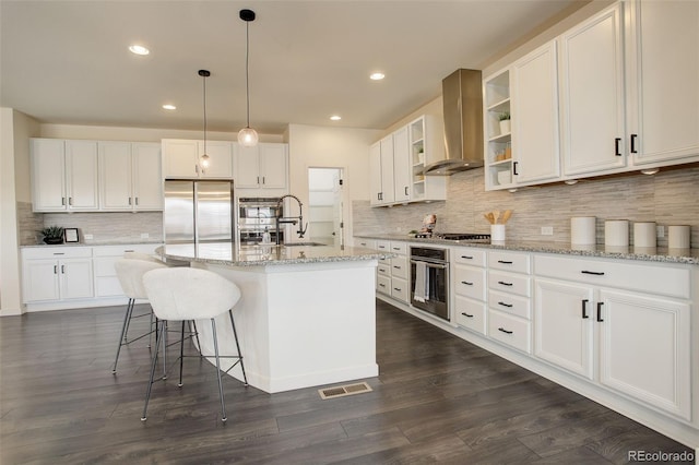 kitchen with wall chimney exhaust hood, stainless steel appliances, an island with sink, decorative light fixtures, and white cabinetry