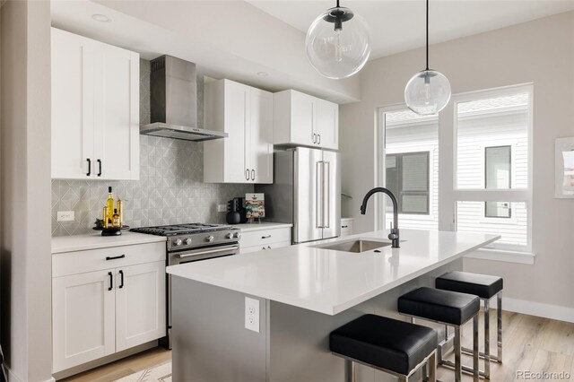 kitchen featuring wall chimney range hood, a sink, light wood-style flooring, appliances with stainless steel finishes, and an island with sink