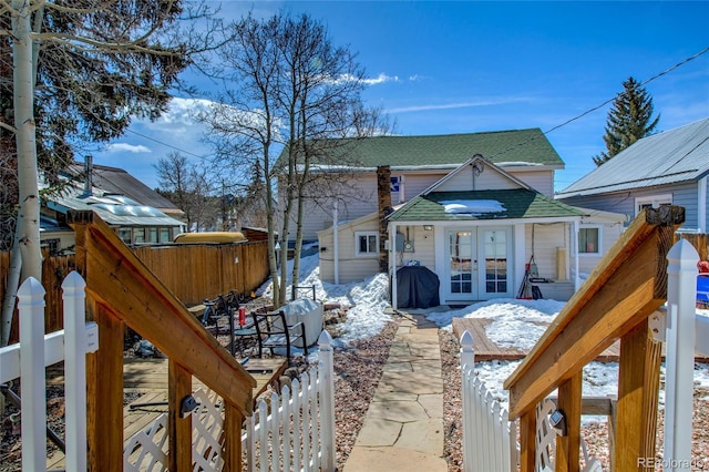 snow covered house featuring french doors, fence, and a shingled roof
