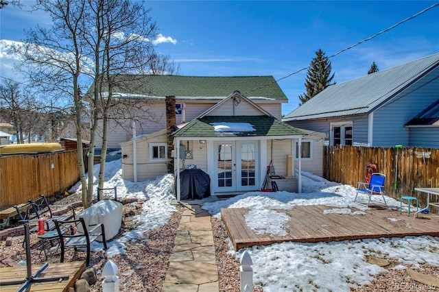snow covered back of property with a shingled roof, french doors, a wooden deck, and fence