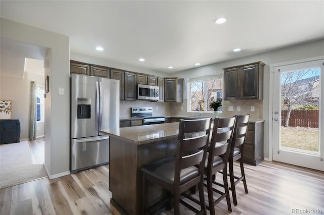 kitchen with tasteful backsplash, dark brown cabinets, a kitchen island, appliances with stainless steel finishes, and light wood-style floors