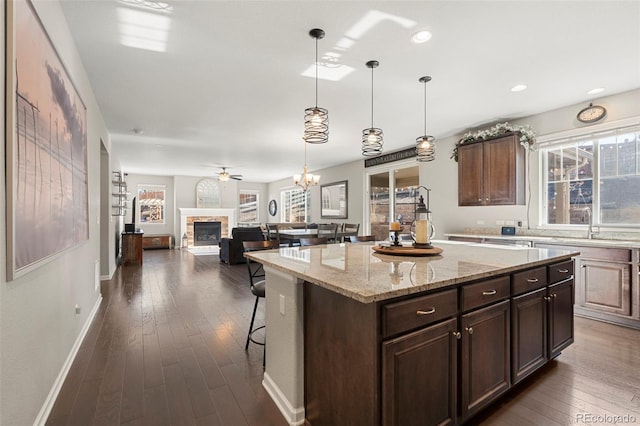 kitchen with light stone counters, a breakfast bar, a fireplace with raised hearth, dark wood-style flooring, and a center island