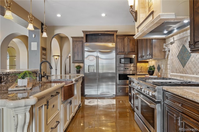 kitchen featuring tasteful backsplash, light stone countertops, a sink, built in appliances, and custom range hood