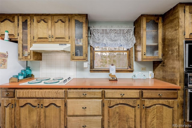 kitchen featuring brown cabinets, backsplash, glass insert cabinets, white electric cooktop, and under cabinet range hood