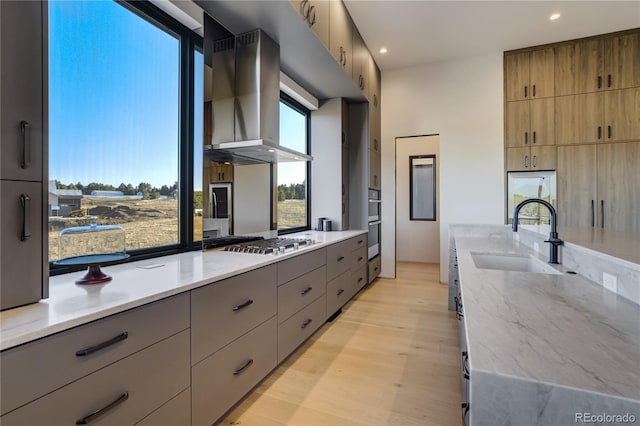 kitchen with wall chimney exhaust hood, light stone countertops, gray cabinets, stainless steel appliances, and a sink