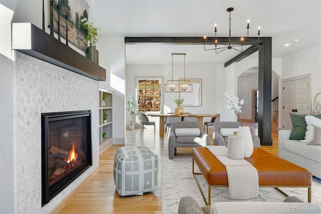 living area with arched walkways, light wood-style floors, a glass covered fireplace, beamed ceiling, and a chandelier